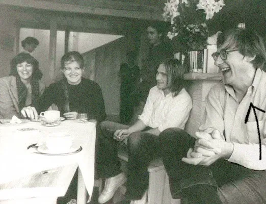 A group of people sits around a table set with teacups and saucers. Christopher Alexander sits on the right, clapping his hands and laughing as if at a hilarious joke.