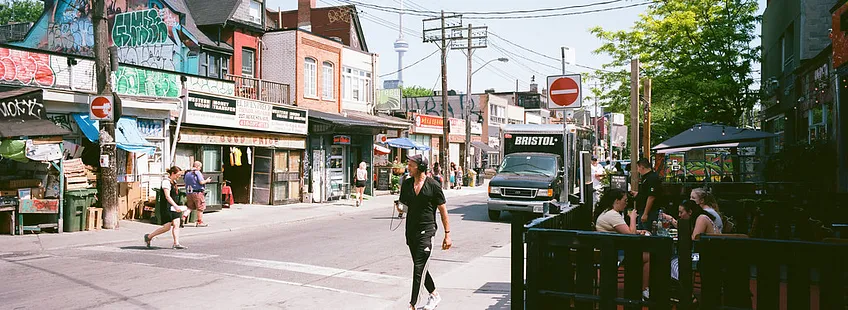 Kensington Market. a street with market stands, people walking around, some sitting outside a cafe. the buildings are small and funky and there is graffiti on the roofs.